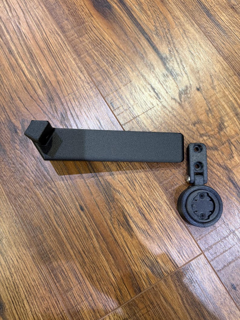 Puede incluir: Soporte negro para ordenador de bicicleta con un brazo rectangular y una base redonda. El soporte est&aacute; hecho de un material negro mate y est&aacute; dise&ntilde;ado para fijarse al manillar de una bicicleta.