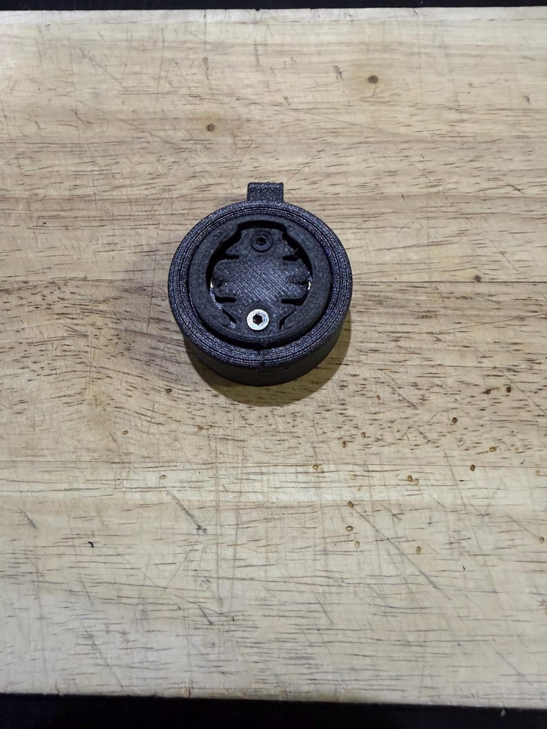 Puede incluir: Soporte negro y circular para ordenador de bicicleta. El soporte tiene una superficie texturizada y un &aacute;rea central dise&ntilde;ada para sujetar un dispositivo. Se ve un peque&ntilde;o tornillo plateado en el centro.