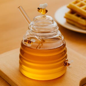 Puede incluir: Un tarro de miel de vidrio transparente con forma de colmena, lleno de miel dorada. El tarro tiene una pajita de vidrio y está decorado con tres pequeñas figuras de abeja. El tarro está sobre una superficie de madera.