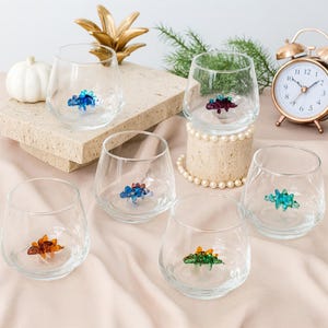 Può includere: Set di sei bicchieri da vino senza stelo trasparenti, ciascuno con una colorata figurina di dinosauro alla base. I dinosauri sono di vari colori, tra cui blu, verde, arancione e rosso. I bicchieri sono disposti su una superficie rosa chiaro.