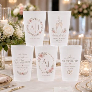 Op de afbeelding: Vijf witte plastic bruiloftbekers met aangepaste tekst en bloemmotieven. De bekers bevatten zinnen als "cheers to love" en "eat, drink, and be married", samen met namen en data. De bekers zijn versierd met roze en groene bloemaccenten.