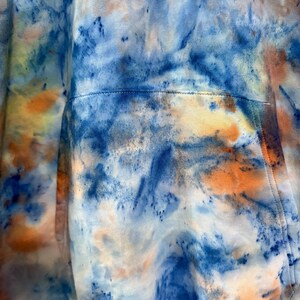 May include: Close-up of a tie-dye hoodie in shades of blue, orange, and white. The fabric has a marbled effect with a pocket detail visible. The hoodie's design features a blend of colors.
