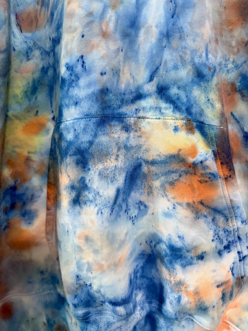 May include: Close-up of a tie-dye hoodie in shades of blue, orange, and white. The fabric has a marbled effect with a pocket detail visible. The hoodie's design features a blend of colors.