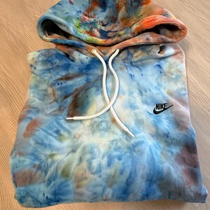 May include: A tie-dye hoodie in shades of blue, orange, and brown. The hoodie features a drawstring hood and the Nike logo on the chest. The garment is made of a soft material.