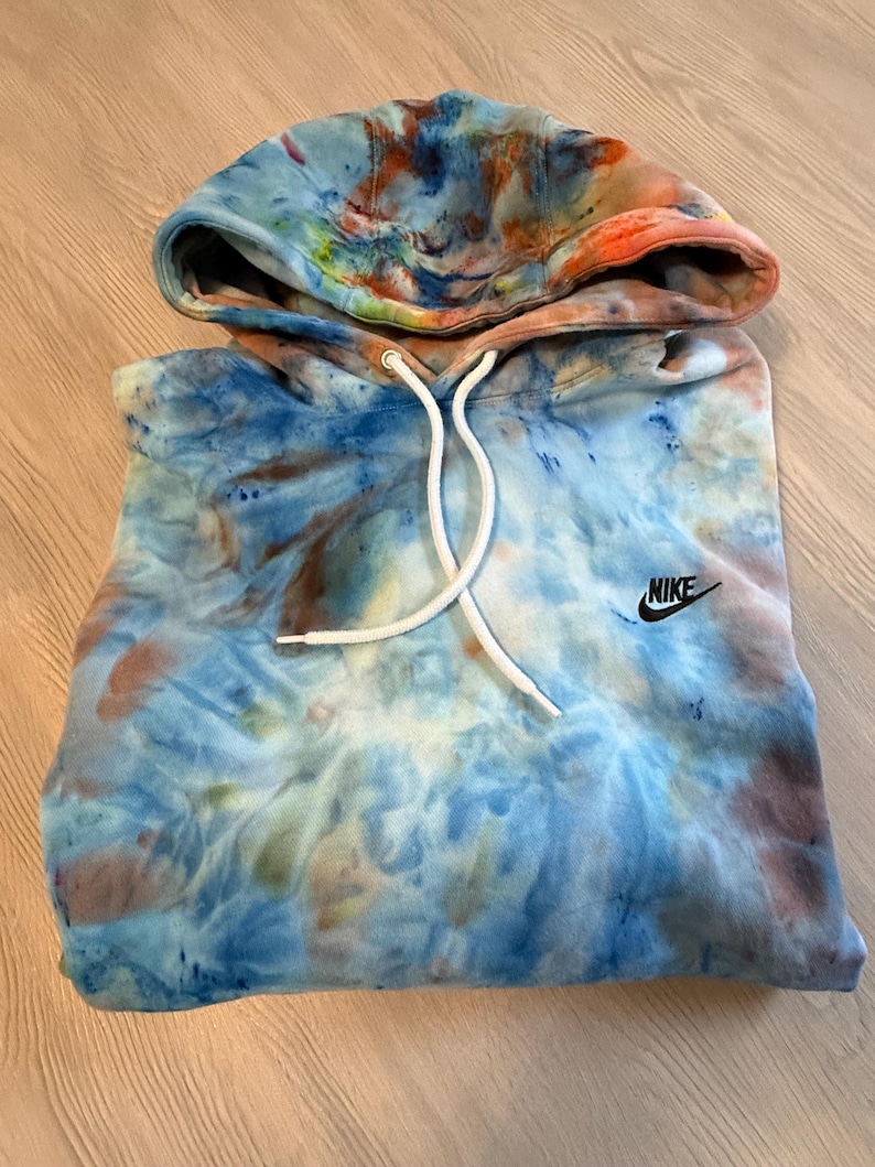 May include: A tie-dye hoodie in shades of blue, orange, and brown. The hoodie features a drawstring hood and the Nike logo on the chest. The garment is made of a soft material.