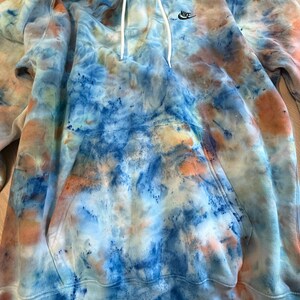 May include: A tie-dye hoodie in shades of blue, orange, and white. The hoodie features a front pocket, a drawstring hood, and the Nike logo on the chest. The garment is made of a soft material.