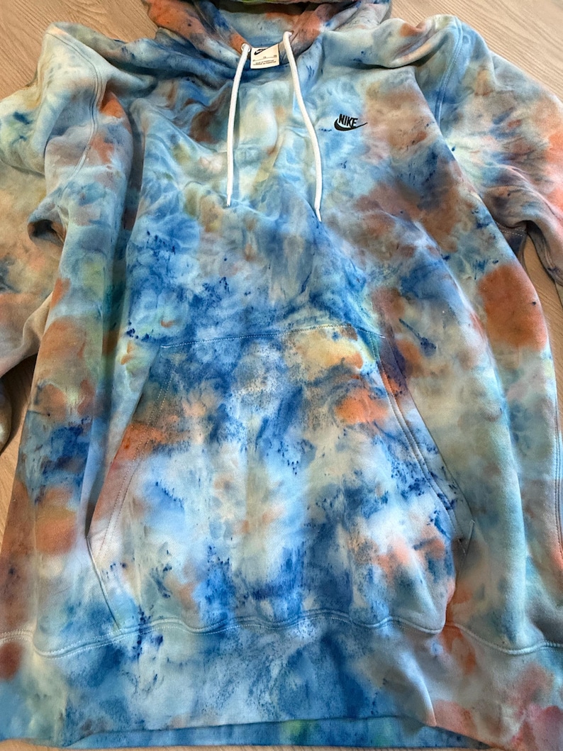 May include: A tie-dye hoodie in shades of blue, orange, and white. The hoodie features a front pocket, a drawstring hood, and the Nike logo on the chest. The garment is made of a soft material.