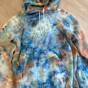 May include: A tie-dye hoodie in shades of blue, orange, and white. The hoodie features a drawstring hood, a front pocket, and the Nike logo on the chest. The garment is made of a soft, comfortable material.