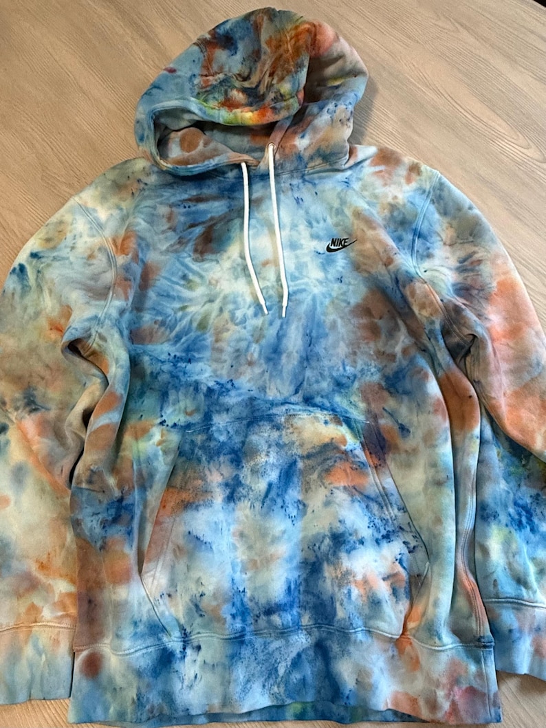May include: A tie-dye hoodie in shades of blue, orange, and white. The hoodie features a drawstring hood, a front pocket, and the Nike logo on the chest. The garment is made of a soft, comfortable material.