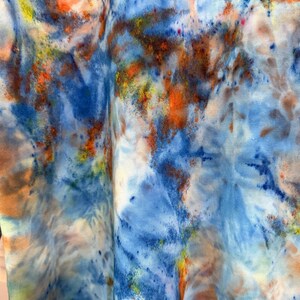 May include: Close-up of a fabric with a tie-dye pattern. The design features a blend of colors, including shades of blue, orange, yellow, and white. The fabric appears to be soft and flowing, with the colors blending and swirling together.