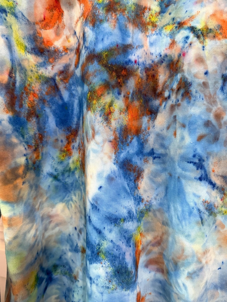 May include: Close-up of a fabric with a tie-dye pattern. The design features a blend of colors, including shades of blue, orange, yellow, and white. The fabric appears to be soft and flowing, with the colors blending and swirling together.