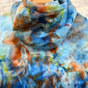 May include: A tie-dye hoodie in shades of blue, orange, and yellow. The hood is up, revealing the interior lining. The fabric has a watercolor-like effect, with the colors blending together. The hoodie is hanging on a wooden hanger.