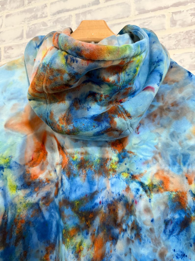 May include: A tie-dye hoodie in shades of blue, orange, and yellow. The hood is up, revealing the interior lining. The fabric has a watercolor-like effect, with the colors blending together. The hoodie is hanging on a wooden hanger.