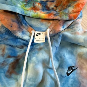 May include: A tie-dye Nike hoodie in shades of blue, orange, and brown. The hoodie features a drawstring hood, a small black Nike logo on the chest, and a white tag with the Nike logo. The hoodie is made in Pakistan.