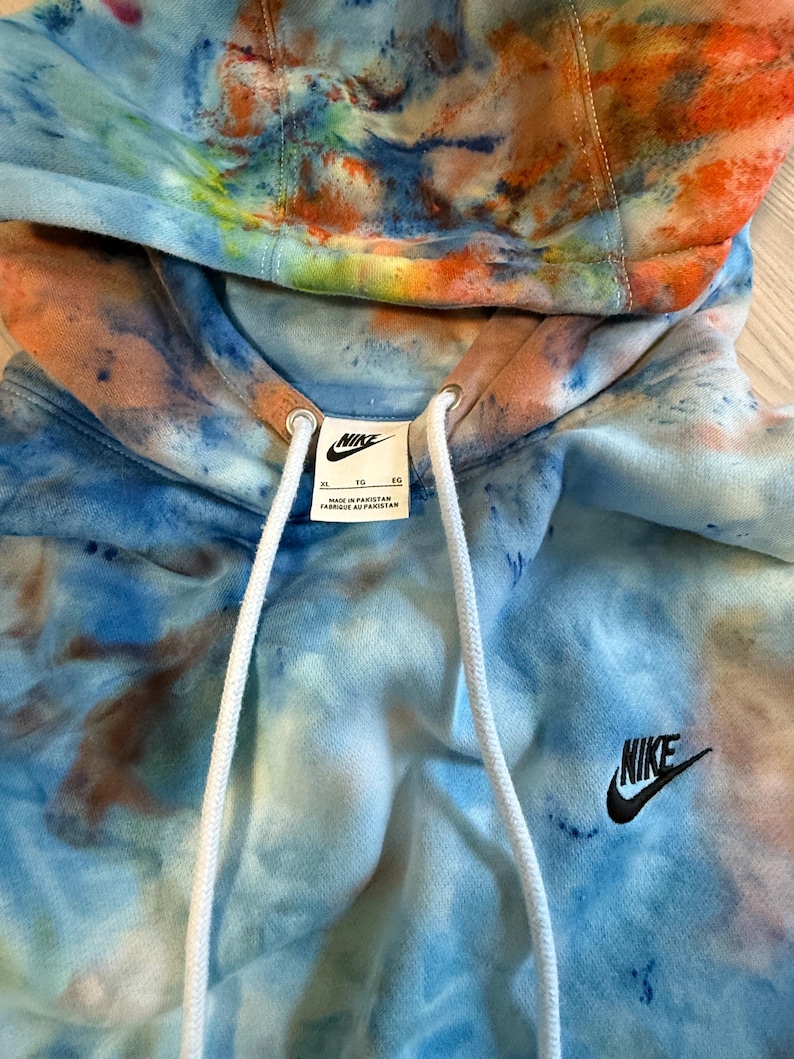 May include: A tie-dye Nike hoodie in shades of blue, orange, and brown. The hoodie features a drawstring hood, a small black Nike logo on the chest, and a white tag with the Nike logo. The hoodie is made in Pakistan.