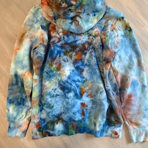 May include: A tie-dye hoodie in shades of blue, orange, and white. The hoodie features a drawstring hood, long sleeves, and ribbed cuffs and hem. The tie-dye pattern is a mix of splatters and swirls.