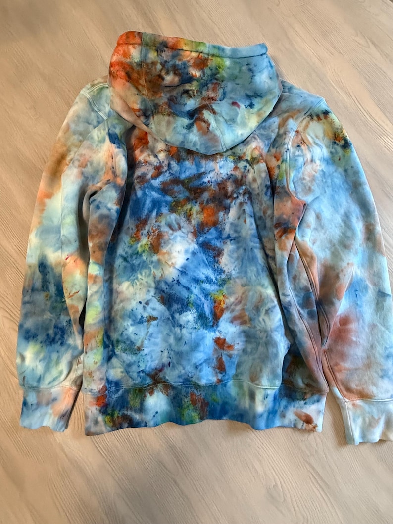 May include: A tie-dye hoodie in shades of blue, orange, and white. The hoodie features a drawstring hood, long sleeves, and ribbed cuffs and hem. The tie-dye pattern is a mix of splatters and swirls.