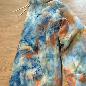 May include: A tie-dye hoodie in shades of blue, orange, and white. The hoodie features a drawstring hood, a front pocket, and the Nike logo on the chest. The garment is made of a soft material.