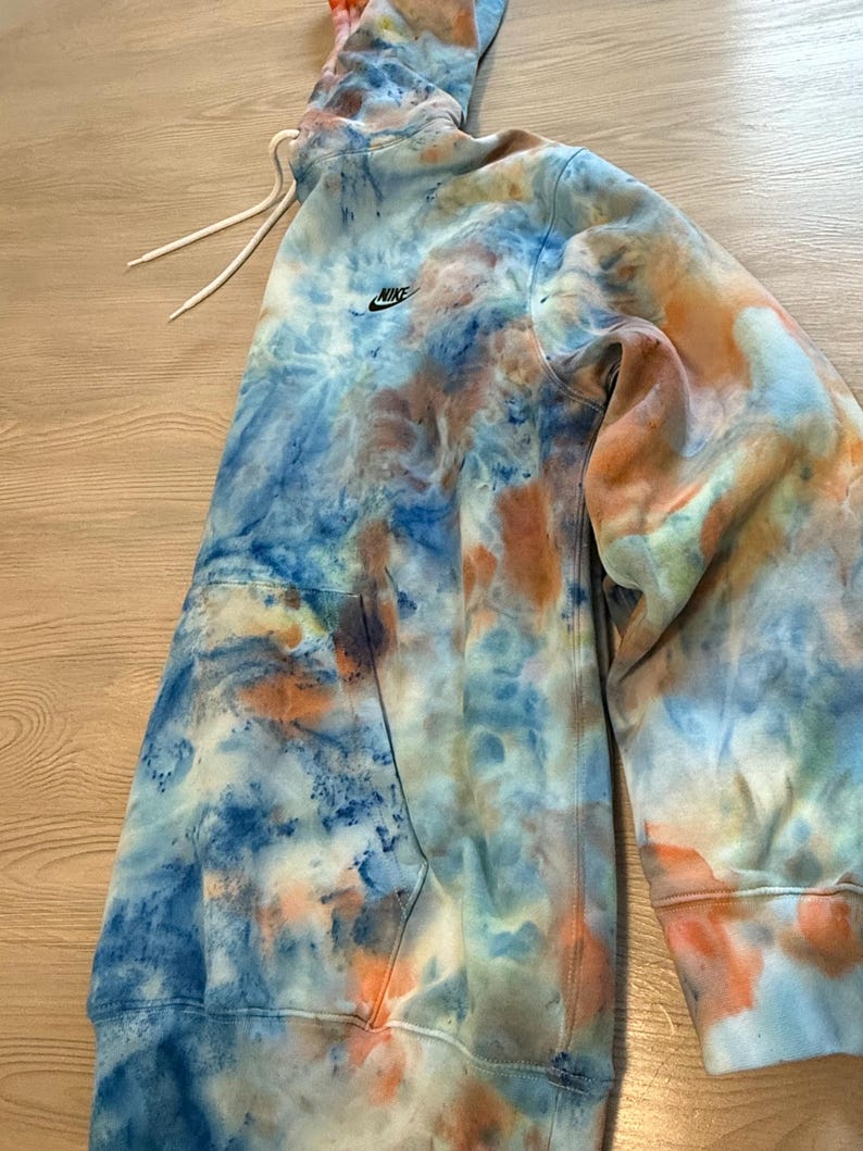 May include: A tie-dye hoodie in shades of blue, orange, and white. The hoodie features a drawstring hood, a front pocket, and the Nike logo on the chest. The garment is made of a soft material.