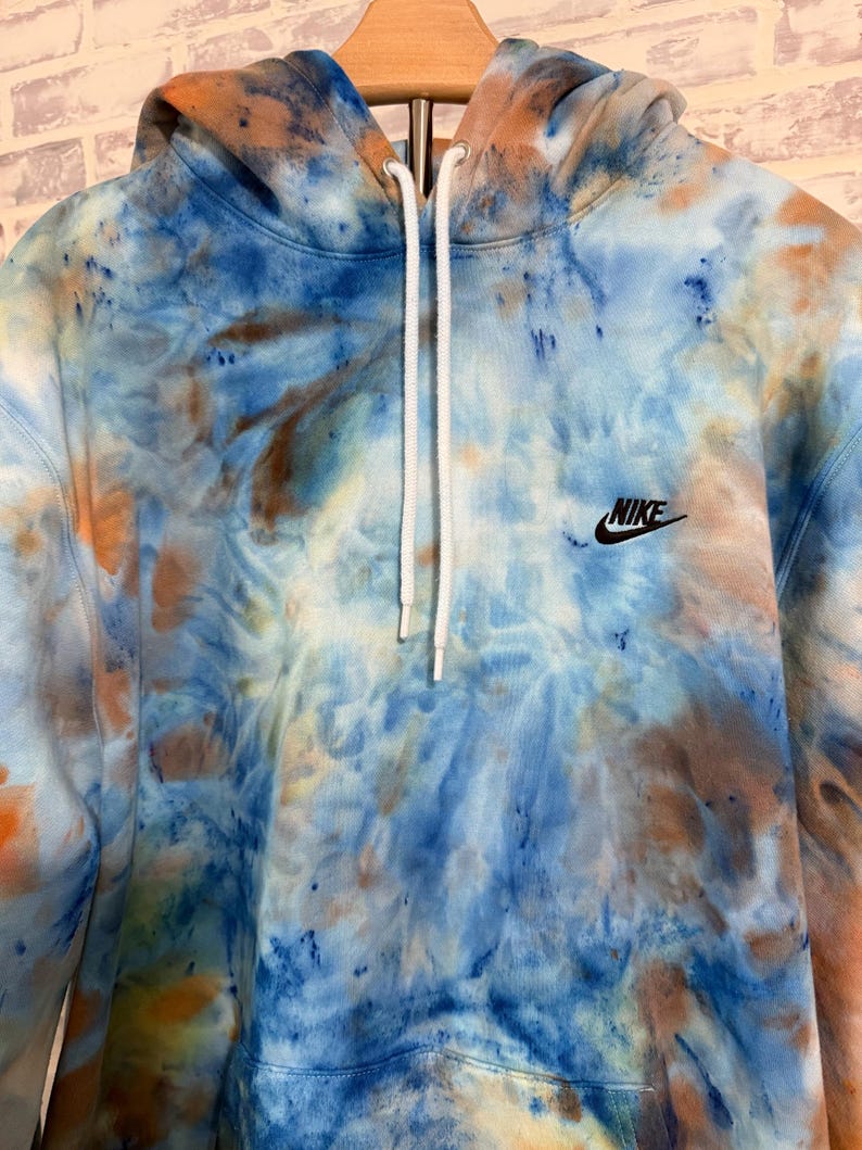 May include: A tie-dye hoodie in shades of blue, orange, and brown. The pullover sweatshirt features a drawstring hood, a front pocket, and the Nike logo embroidered on the chest. The garment is displayed on a wooden hanger.