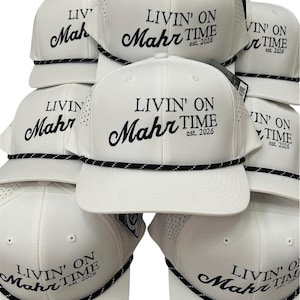 Peut inclure: Casquettes de baseball blanches avec le texte "LIVIN' ON Mahr TIME est. 2026" brodé en noir. Les casquettes ont un détail de corde noire et blanche le long du bord et de petits trous de ventilation. Les casquettes sont empilées.