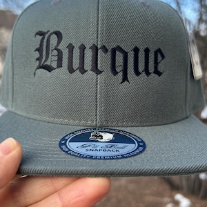 May include: A gray snapback hat with the word "Burque" in black gothic lettering. The hat has a flat brim and a "Pit Bull Snapback" patch. The hat is made of a textured fabric.