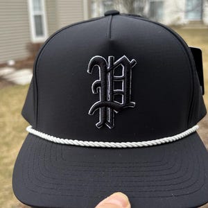 Könnte beinhalten: Schwarze Baseballkappe mit weißem Seildetail und einem weiß gestickten Buchstaben "P" auf der Vorderseite. Die Kappe hat einen gebogenen Schirm und eine strukturierte Passform. Ein Etikett ist an der Seite befestigt.