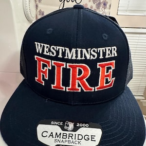 May include: A navy blue snapback cap with the words "WESTMINSTER FIRE" in white and red lettering. The cap has a black mesh back and a curved brim. A tag on the brim reads "CAMBRIDGE SNAPBACK" and "PB107".