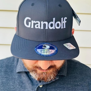 Puede incluir: Gorra snapback gris antracita con la palabra "Grandolf" bordada en blanco. La gorra tiene una visera plana y una pegatina "Pit Bull Snapback". La persona de la imagen lleva un polo gris.