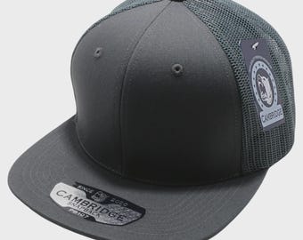 Op de afbeelding: Een antracietgrijze snapback pet met een mesh achterkant en een platte klep. De pet heeft de tekst "CAMBRIDGE SNAPBACK" op een label en op de klep. De pet heeft een gestructureerde kroon en een knoop aan de bovenkant.