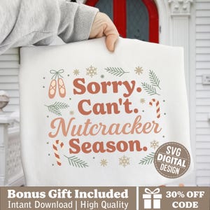 May include: A white tote bag with the text "Sorry. Can't. Nutcracker Season." in shades of brown. The design includes ballet shoes, snowflakes, candy canes, and sprigs of greenery. A digital design logo is in the lower right corner.