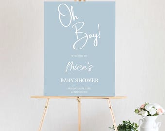 Pode incluir: Uma placa azul clara em um cavalete de madeira com as palavras "Oh Boy!" em elegante escrita branca. A placa dá as boas-vindas aos convidados para o chá de bebê de Mica, com a data e o local na parte inferior. Um vaso de flores está por perto.