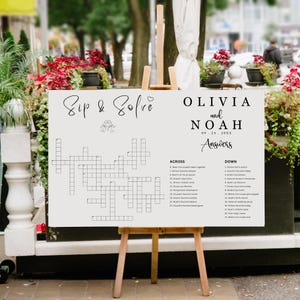 May include: A cream-colored wedding crossword puzzle sign on a wooden easel. The sign reads "Sip & Solve" at the top, with the names "Olivia and Noah" and the date "09.24.20XX" below. The crossword has clues listed on the sides.