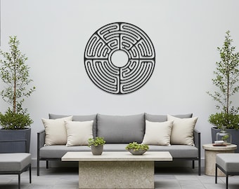 Op de afbeelding: Een zwart metalen labyrint muurkunstwerk is bevestigd aan een witte muur boven een grijze buitenzitbank. Het cirkelvormige ontwerp heeft een doolhofpatroon. De buitenomgeving omvat een stenen salontafel, potplanten en een grijs terras.