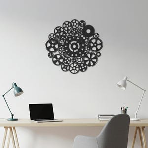 Puede incluir: Una pieza de arte mural de metal negro que presenta una compleja disposición de engranajes de varios tamaños y formas. Los engranajes están agrupados en un diseño circular, creando una estética mecánica e industrial. La obra de arte está montada en una pared blanca.