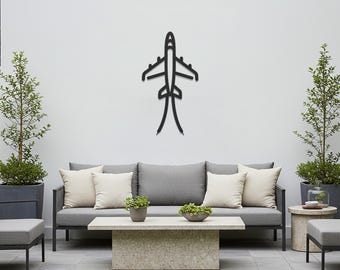 May include: A black metal airplane wall art decor is mounted on a white wall above a gray outdoor sofa with cream-colored pillows. The outdoor setting includes a stone coffee table, side tables, and potted plants.