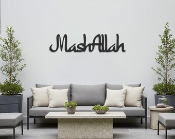 May include: A black metal wall art piece that reads "MashAllah" in elegant cursive script. The artwork is mounted on a white wall above a gray outdoor sofa with cream-colored pillows. The scene includes a stone coffee table and potted plants.