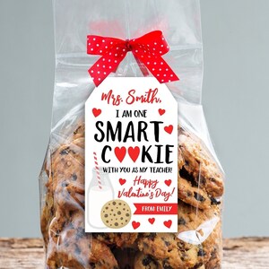 May include: A clear cellophane bag filled with chocolate chip cookies, tied with a red polka dot ribbon. A white tag with red hearts and text reads "Mrs. Smith, I AM ONE SMART COOKIE with you as my teacher! Happy Valentine's Day! From: Emily."