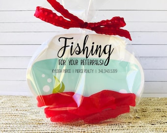 May include: A clear plastic bag filled with red candy shaped like fish. The bag is tied with a red ribbon and has a white tag with the text "Fishing for your referrals! KYLEIGH PIERCE | PIERCE REALTY | 341.343.5319"