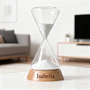May include: Clear glass hourglass with white sand, set on a wooden base. The base is engraved with the name "Isabella". The hourglass is a decorative item, likely for home decor or as a gift.