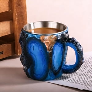 May include: A blue and brown coffee mug with a stainless steel rim. The mug has a unique, textured design resembling a geode. The mug is filled with a brown liquid, likely coffee or tea.