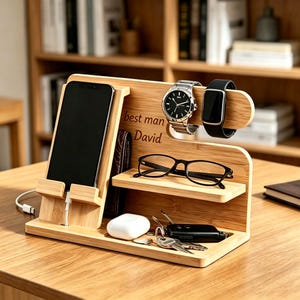 May include: A light-colored bamboo wood docking station featuring a vertical smartphone stand with a charging cable slot, a horizontal shelf for eyeglasses, and a base tray for accessories. The wood is engraved with the text best man David.