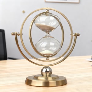 May include: An hourglass with a clear glass bulb filled with sand, set in a gold-colored metal frame. The hourglass is supported by a circular base and a spherical accent. The design features a double-loop frame, creating a decorative and functional timekeeping device.