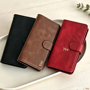 Puede incluir: Tres fundas rectangulares para teléfono tipo cartera en negro, marrón y rojo. Las fundas marrón y roja tienen una lengüeta de cierre magnético y el nombre Yee grabado en letras doradas en la cubierta frontal. Acabado mate y liso.
