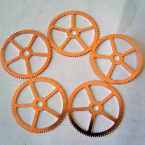 May include: Five copper-colored watch gears arranged in a circular pattern. Each gear has a central hole and five spokes connecting to the toothed outer rim. The gears are evenly spaced against a light background.