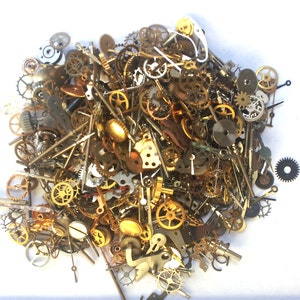 May include: A large pile of assorted gold and silver clock gears, springs, and other watch parts. The parts are scattered on a white surface.