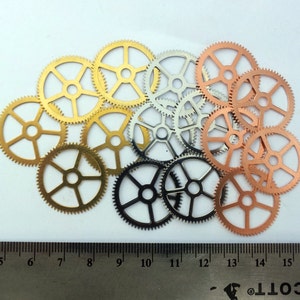 May include: A collection of 13 metal gears in various sizes and colors, including gold, silver, and copper. The gears are arranged in a random pattern on a white surface.