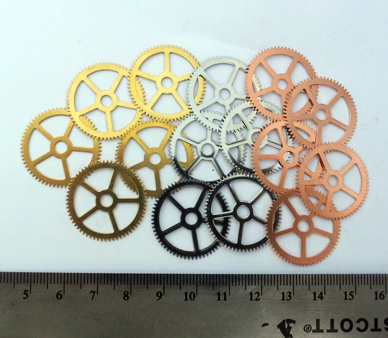 May include: A collection of 13 metal gears in various sizes and colors, including gold, silver, and copper. The gears are arranged in a random pattern on a white surface.