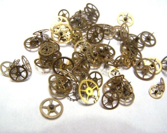 May include: A collection of gold-toned metal gears of various sizes, scattered on a white surface.