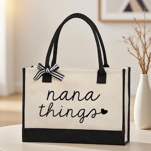 Puede incluir: Bolso tote color crema con detalles y asas negras. Presenta la frase "nana things" en cursiva, con un pequeño corazón. Un lazo a rayas adorna la esquina superior izquierda. Diseñado para transportar objetos.